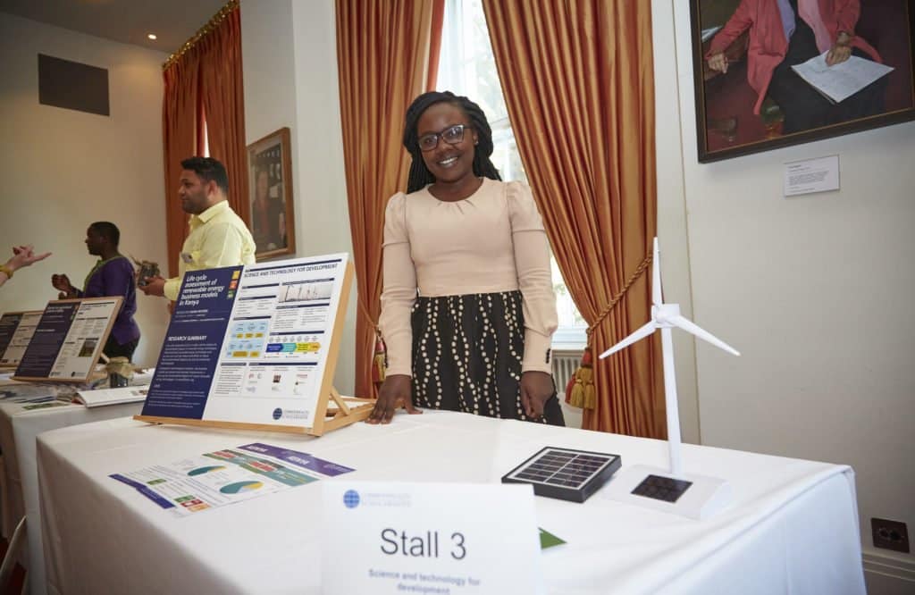  Rose Shikuri Munyendo Okilo, a Commonwealth Scholar from Kenya and an exhibitor, demonstrates the impact of her research. Photo includes small scale models of a wind turbine and solar panels