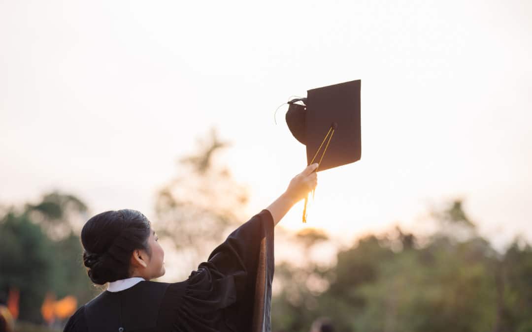 A woman dressed in graduate attire has lifted her graduate hat towards the sky