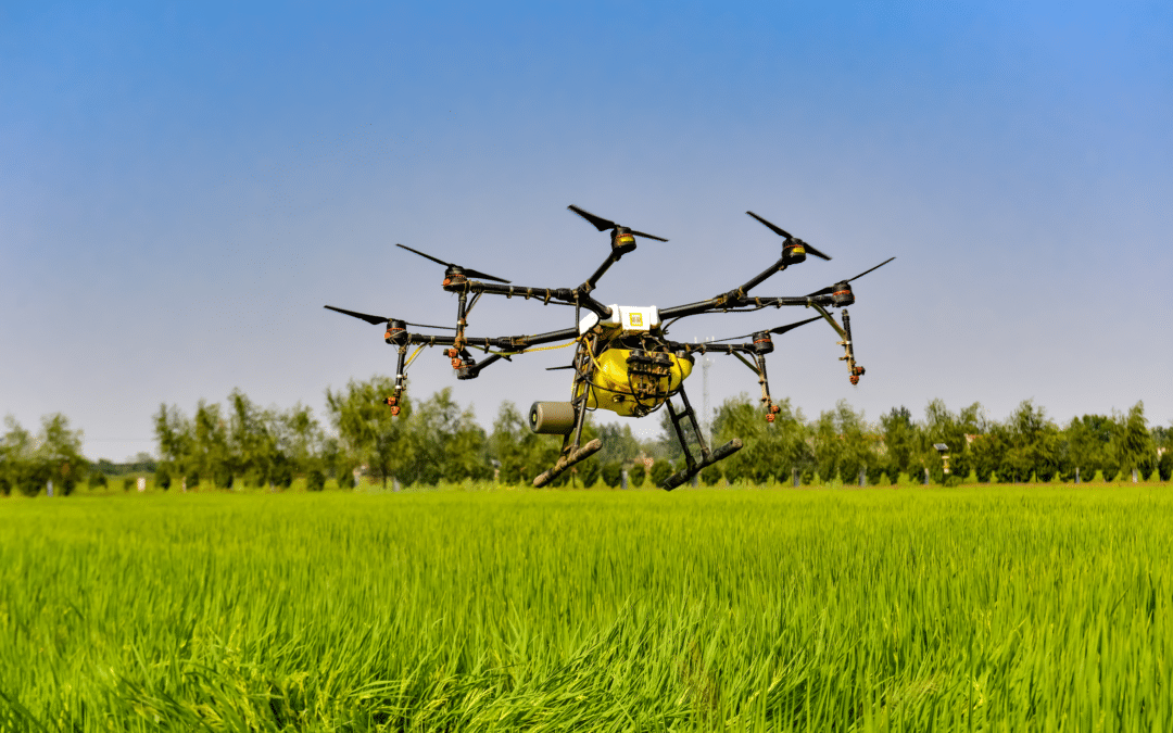 Agricultural drone flying over crop field with trees and sky in background.