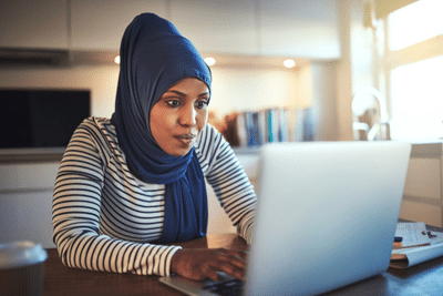 Young woman wearing a hijab sitting at her kitchen table using a laptop.