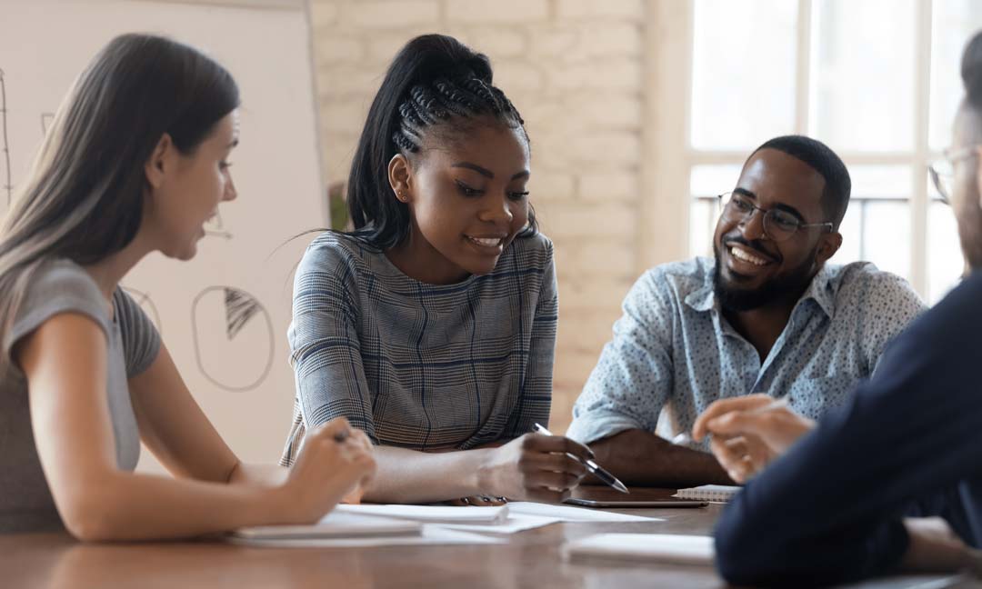 Diverse group of people sitting at a table discussing ideas and making notes, with flipchart in background.